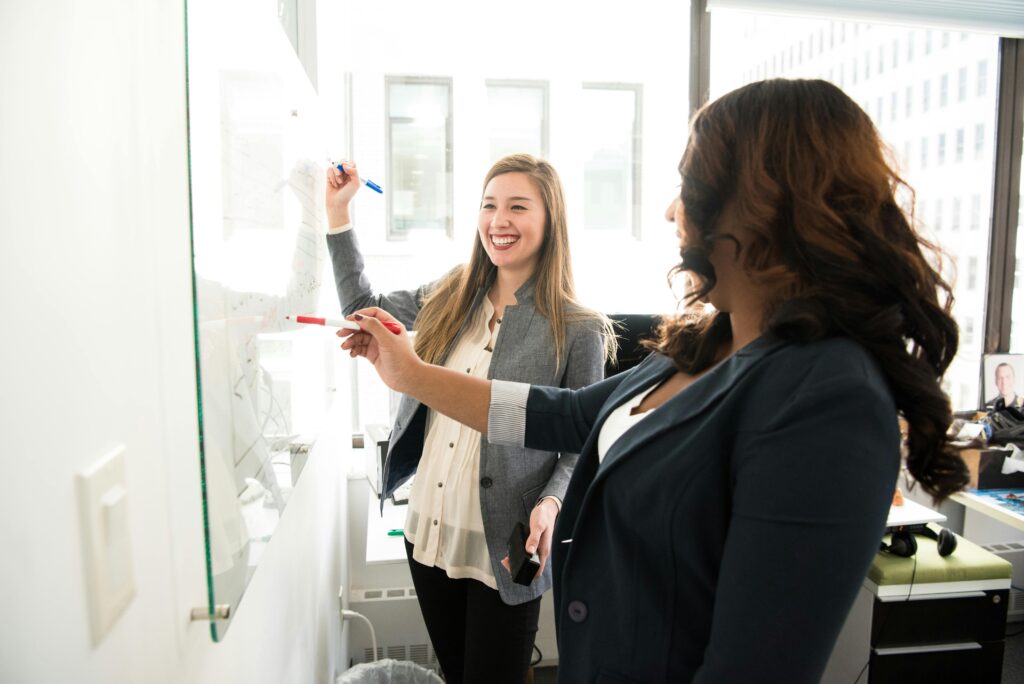 Two business professionals collaborating at a glass whiteboard, discussing ideas and strategy in a modern office environment.
